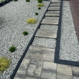 Ułożona ścieżka z płyt betonowych prowadząca przez rabatę z jasnego żwiru i niskich roślin, obrzeża z kostki, widok z góry.