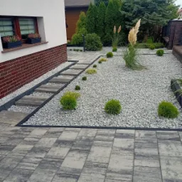 Ogród z szarymi kamieniami, ozdobnymi trawami i kulistymi krzewami, ścieżka z płyt na podwyższeniu, kostka brukowa w tle, donice z kwiatami w oknie domu.