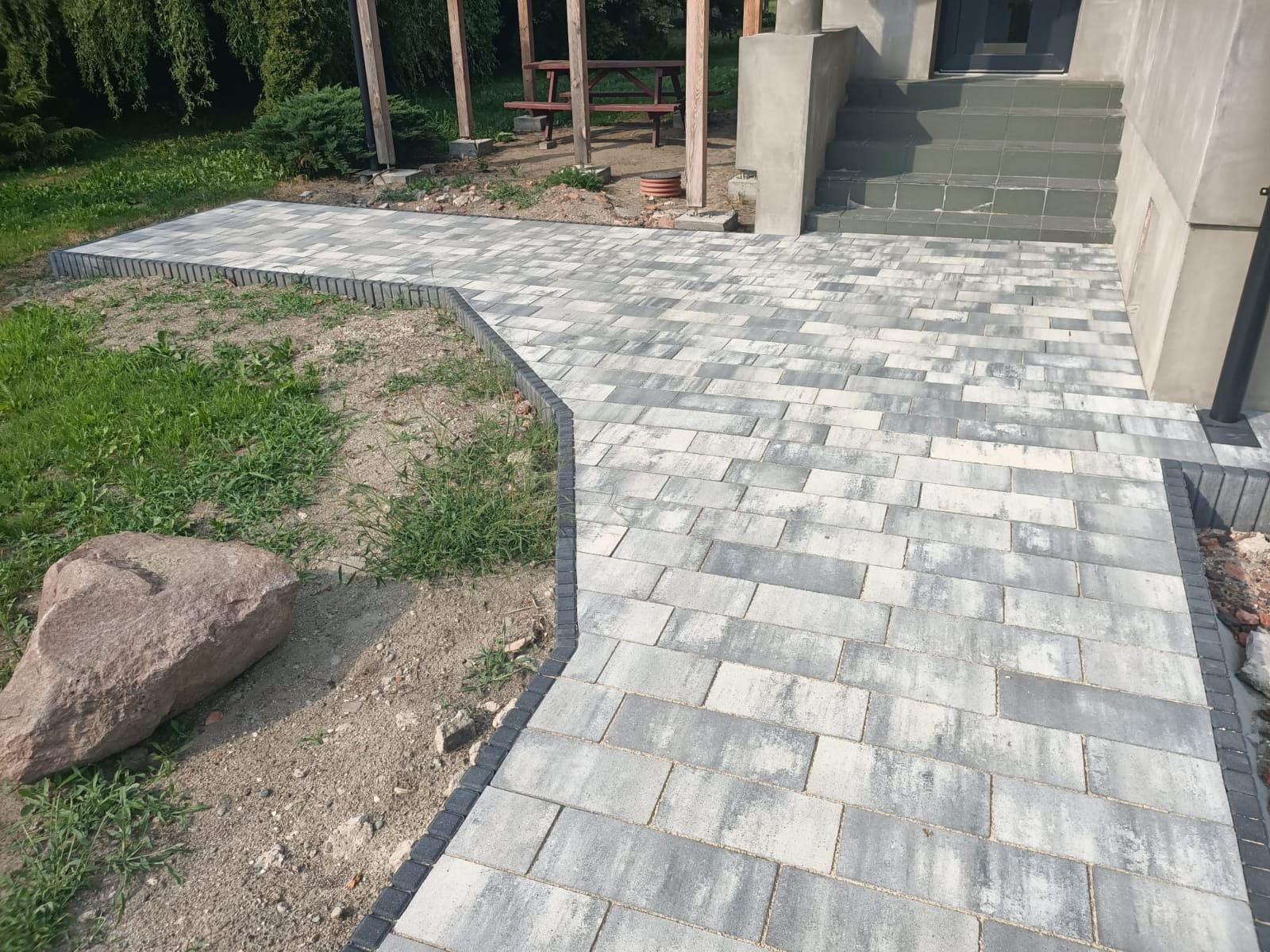 Ścieżka z jasnoszarej kostki brukowej prowadząca do schodów wejściowych, obrzeże z ciemnej kostki, widoczny fragment trawnika z kamieniem.