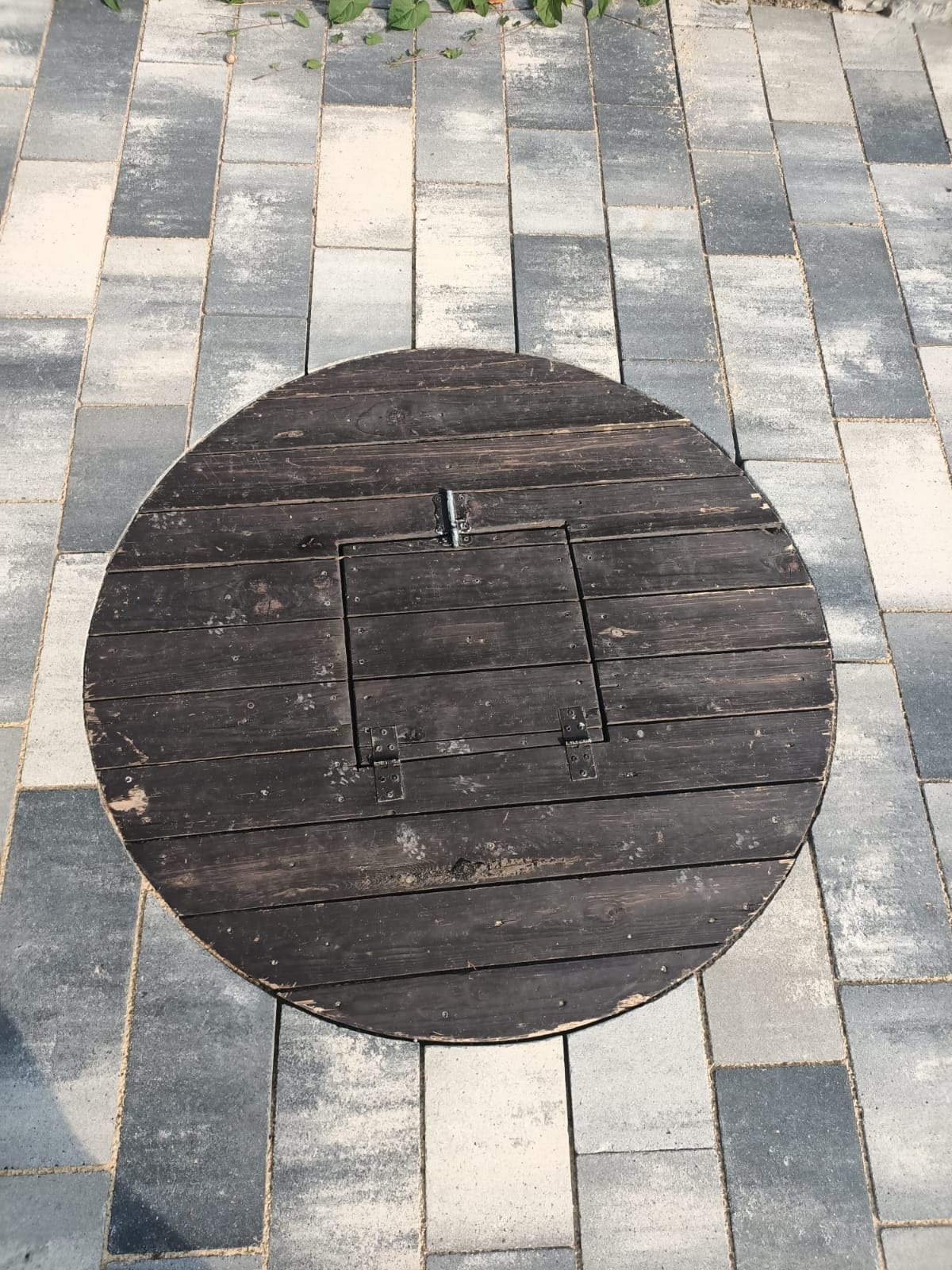 Okrągły, ciemny, drewniany dekiel z klapką, umieszczony na szaro-białej kostce brukowej.