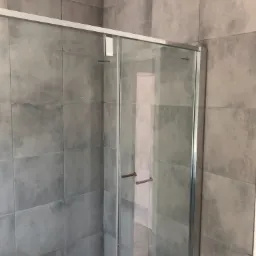 Szklana kabina prysznicowa z srebrnymi profilami na tle ściany wyłożonej szarymi, kwadratowymi płytkami o teksturze betonu.