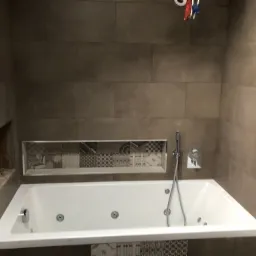 Wykończona łazienka z wanną jacuzzi, obłożona płytkami w odcieniach szarości i beżu, z dekoracyjną wnęką nad wanną wyłożoną mozaiką o zróżnicowanych wzorach.