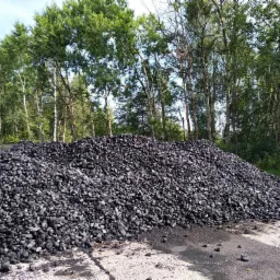 Góra czarnego węgla kamiennego na betonowym placu składowym, w tle drzewa liściaste pod błękitnym niebem.