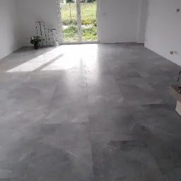 Wykończona posadzka z szarej, polerowanej płyty w jasnym, pustym pomieszczeniu z kominkiem i dużym oknem balkonowym.