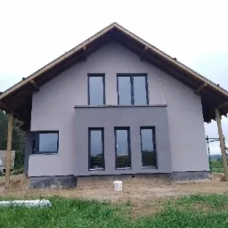 Dom jednorodzinny po nałożeniu szarego tynku elewacyjnego, widoczne czarne ramy okienne i drewniana konstrukcja dachu, teren wokół budynku częściowo zagospodarowany.