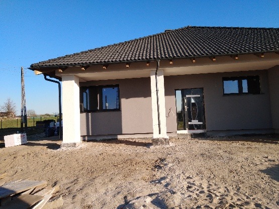 Surowa elewacja domu jednorodzinnego w trakcie prac wykończeniowych, widoczne białe kolumny podtrzymujące zadaszenie i niezabezpieczone fundamenty.