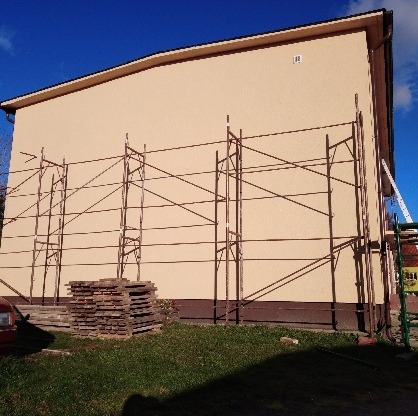 Ocieplona elewacja budynku z beżowym tynkiem, widoczne metalowe rusztowania i stos desek u podnóża ściany, trawnik z przodu.