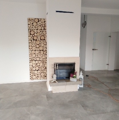 Wnętrze salonu z kominkiem obudowanym jasnym kamieniem, dekoracją z drewna na ścianie, szarymi płytkami podłogowymi i przeszklonymi drzwiami w tle.