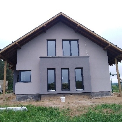 Dom jednorodzinny po nałożeniu szarego tynku elewacyjnego, widoczne czarne ramy okienne i drewniana konstrukcja dachu, teren wokół budynku częściowo zagospodarowany.