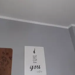 Pomieszczenie z szarą ścianą, białą listwą przysufitową, plakatem z napisem i drewnianą dekoracją. Ściana pomalowana na jednolity, matowy kolor.