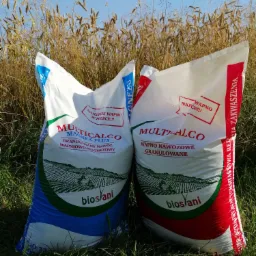 Dwa worki z granulowanym wapnem nawozowym leżą na trawie na tle wysokich traw i błękitnego nieba.