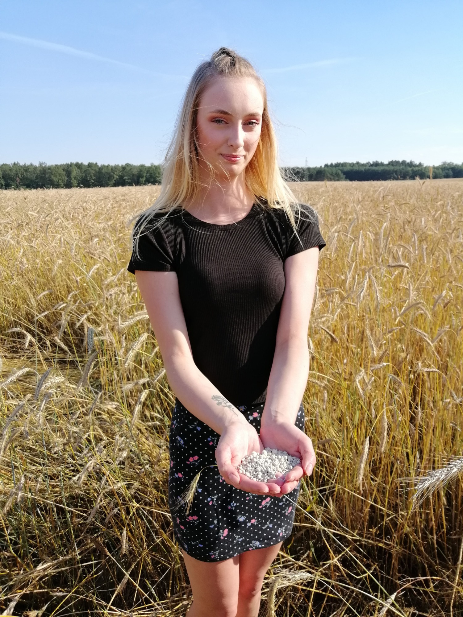 Blondynka trzymająca nawóz mineralny na tle pola zboża w słoneczny dzień.