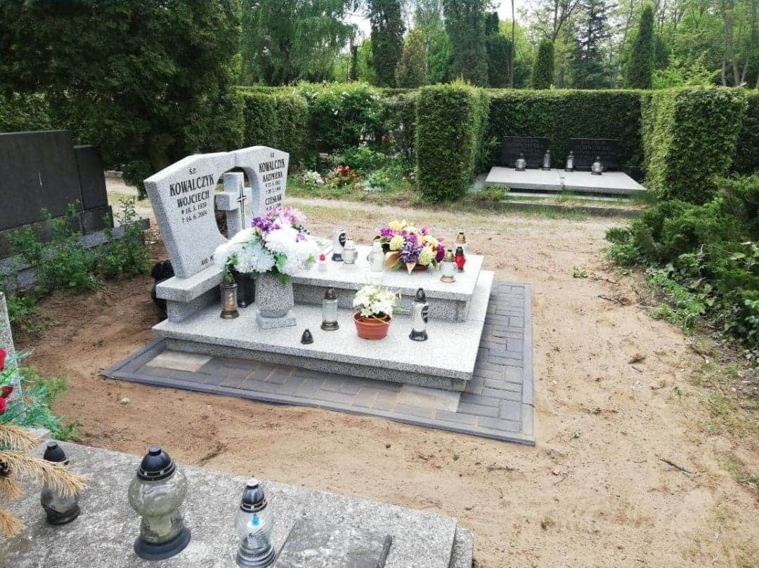 Jasny, granitowy nagrobek z kwiatami i zniczami na cmentarzu, otoczony kostką brukową i zielenią.