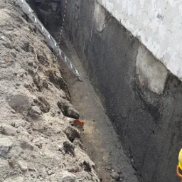 Wykop fundamentów z odsłoniętą ścianą, widoczna drabina oparta o ścianę wykopu, pryzma ziemi i fragment pracownika w żółtym kasku.