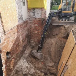 Żółta koparka wykonuje wykop wzdłuż ceglanej ściany fundamentowej zabezpieczonej płytami OSB, widoczne odsłonięte kable i rury.