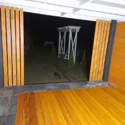 Drewniana altana z widokiem na huśtawkę ogrodową w nocy, widoczne narzędzia i deski podłogowe w trakcie montażu.