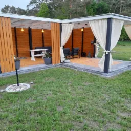 Nowoczesna altana ogrodowa z drewnianymi ścianami, zasłonami w kolorze ecru, oświetleniem punktowym i meblami wypoczynkowymi, otoczona kostką brukową i trawnikiem.