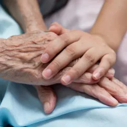 Dłoń osoby młodej spoczywa na pomarszczonej dłoni osoby starszej, leżącej na błękitnym materiale. Ujęcie z bliska, skupione na dłoniach.