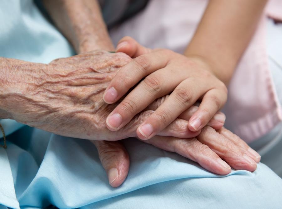Dłoń osoby młodej spoczywa na pomarszczonej dłoni osoby starszej, leżącej na błękitnym materiale. Ujęcie z bliska, skupione na dłoniach.