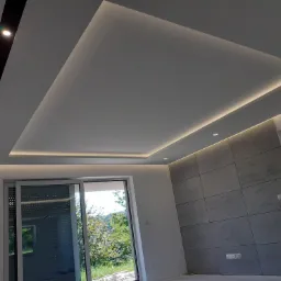 Nowoczesny sufit podwieszany z oświetleniem LED w wykończonym wnętrzu, widoczne okno przesuwne z widokiem na zieleń.