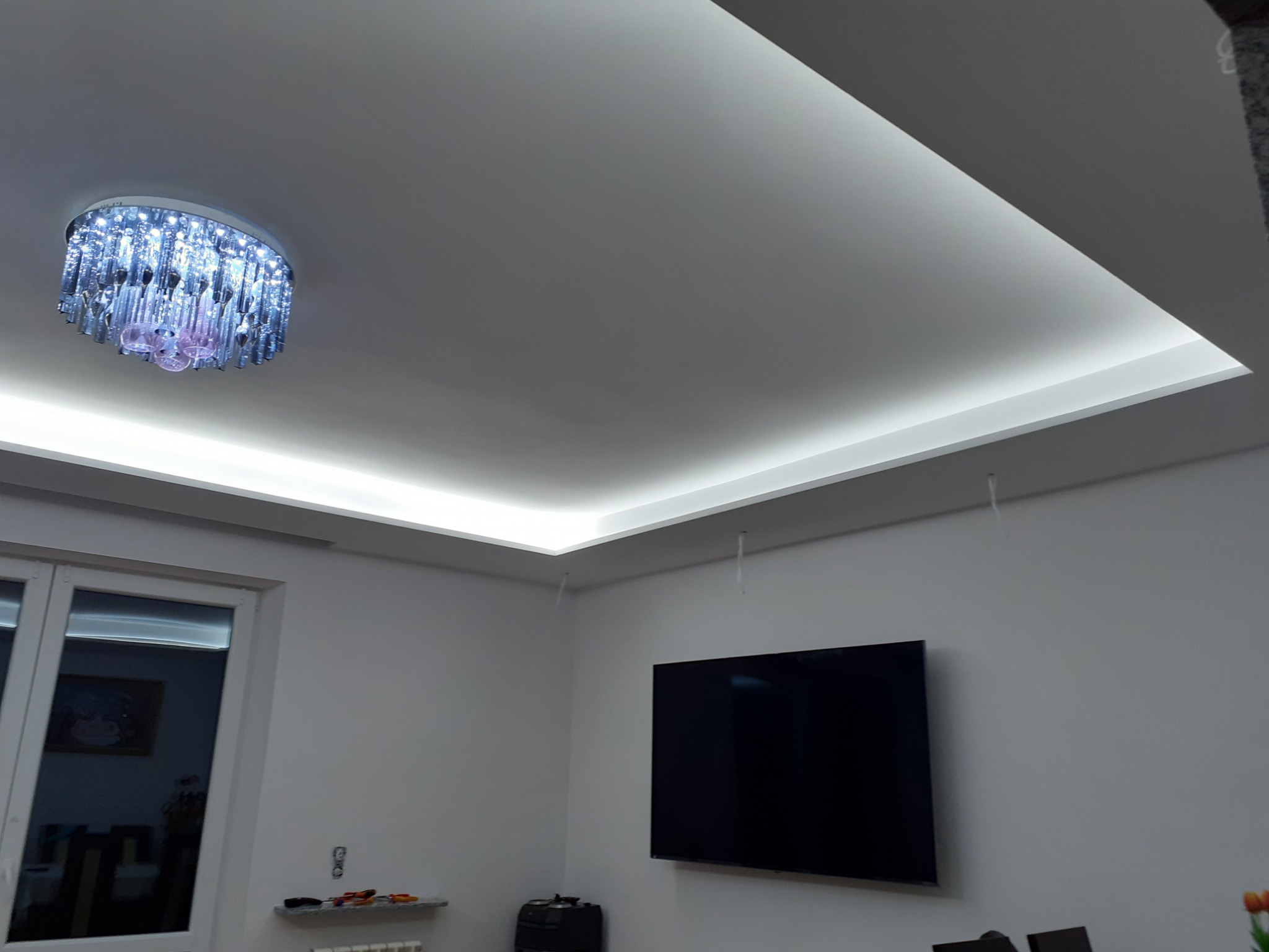 Nowoczesny salon z podwieszanym sufitem, oświetleniem LED i kryształowym żyrandolem, widoczny telewizor na ścianie i okno.