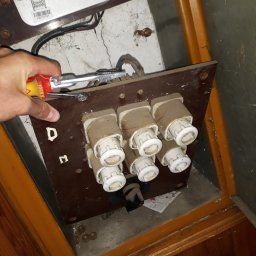 Punktstyku.pl - Usługi Elektryczne - Stara tablica bezpiecznikowa typu Dm z ceramicznymi wkładkami topikowymi, częściowo zdemontowana, widoczna ręka elektryka z narzędziem.