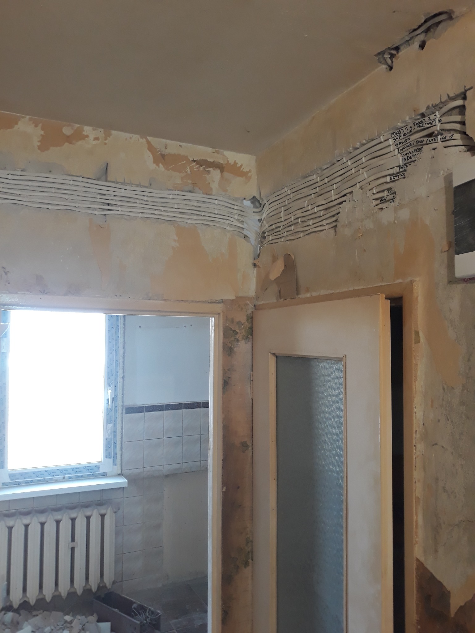 Przedpokój w bloku z widocznymi przewodami elektrycznymi ułożonymi w bruzdach na ścianie, przygotowanymi do tynkowania. Widoczne napisy na ścianie opisujące przeznaczenie obwodów.