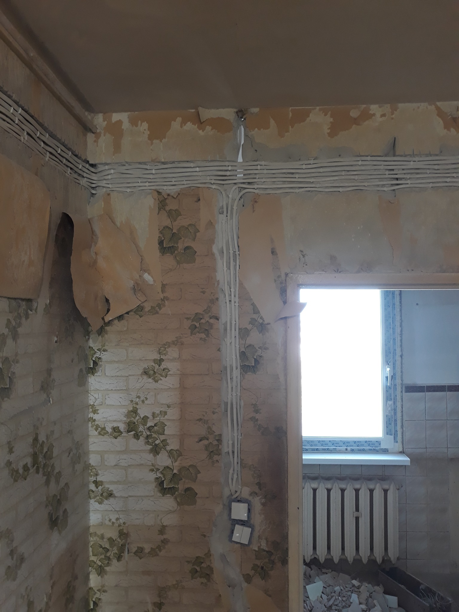 Przedpokój w bloku z widocznymi przewodami elektrycznymi na ścianach przed remontem. Fragmenty tapet odklejone, widoczne puszki pod włączniki.