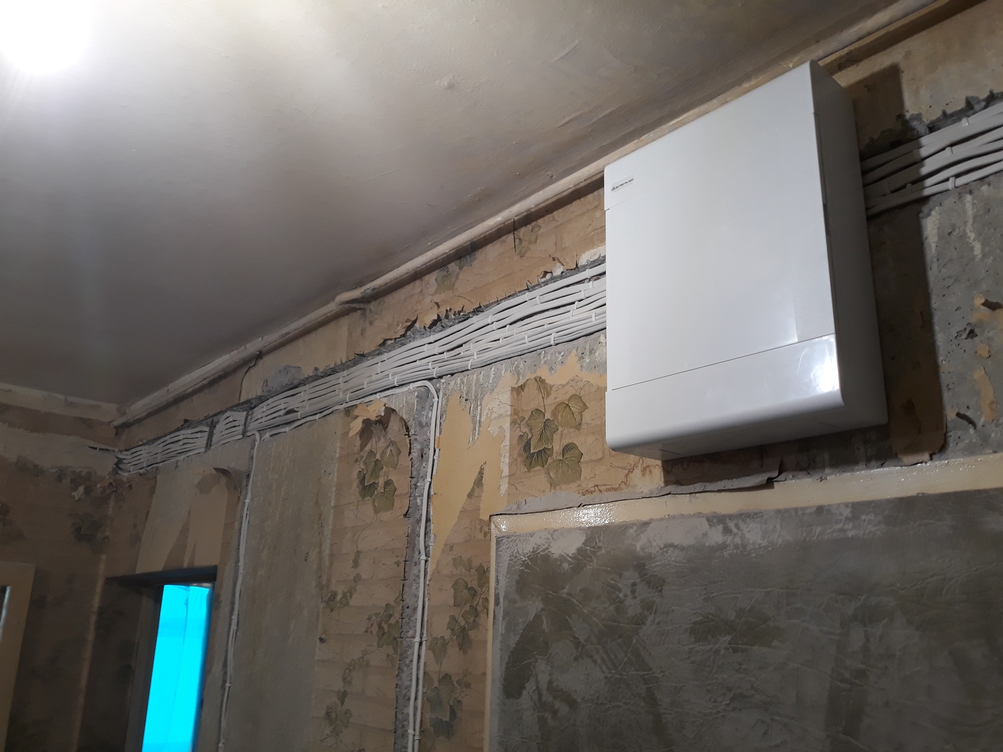 Biały bojler elektryczny zamontowany na ścianie z widocznymi przewodami w listwach. Ściana w trakcie remontu, z fragmentami starej tapety i nowej gładzi.