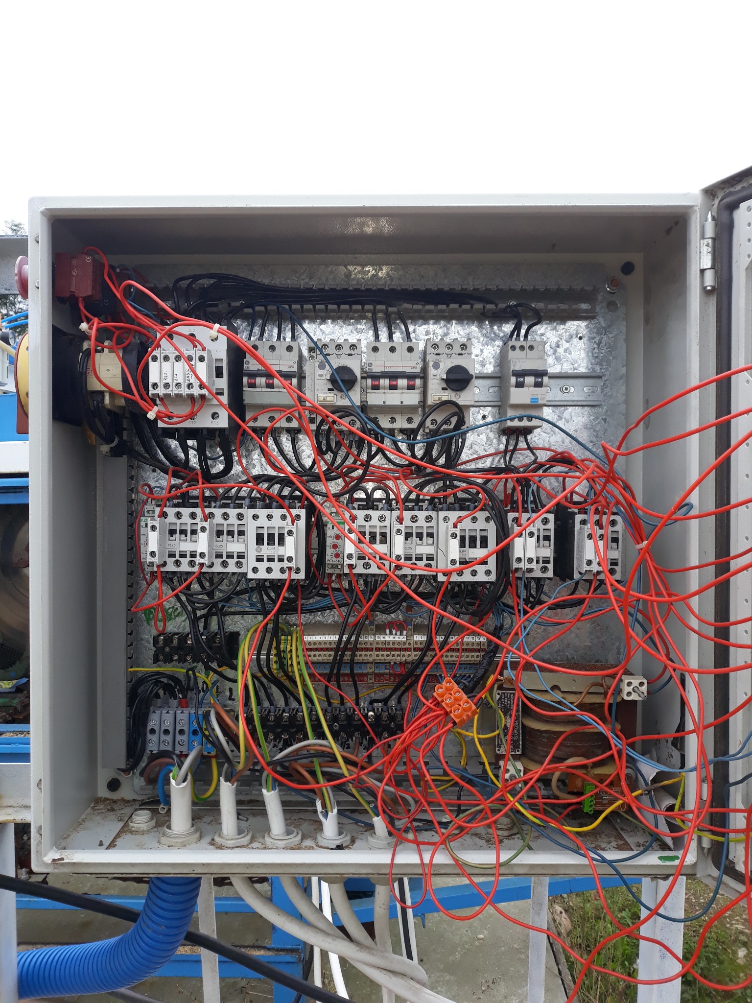 Otwarta skrzynka elektryczna z licznymi przewodami, bezpiecznikami i elementami sterowania. Widoczne kable w kolorach czerwonym, żółtym i niebieskim.