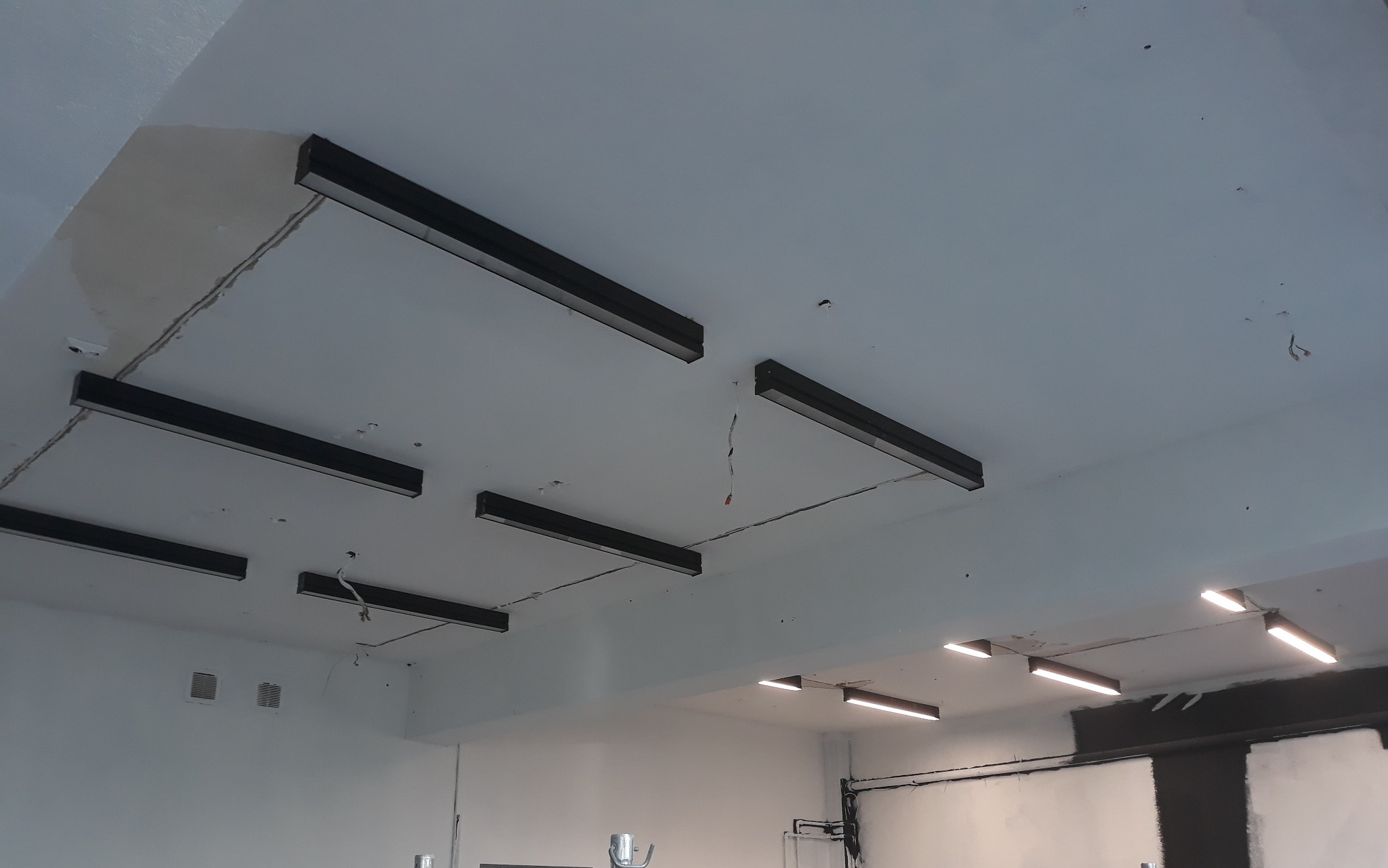 Zamontowane, czarne, podłużne lampy sufitowe w pomieszczeniu z białymi ścianami. Widoczne przewody i niedoskonałości sufitu. Oświetlenie w stylu industrialnym.