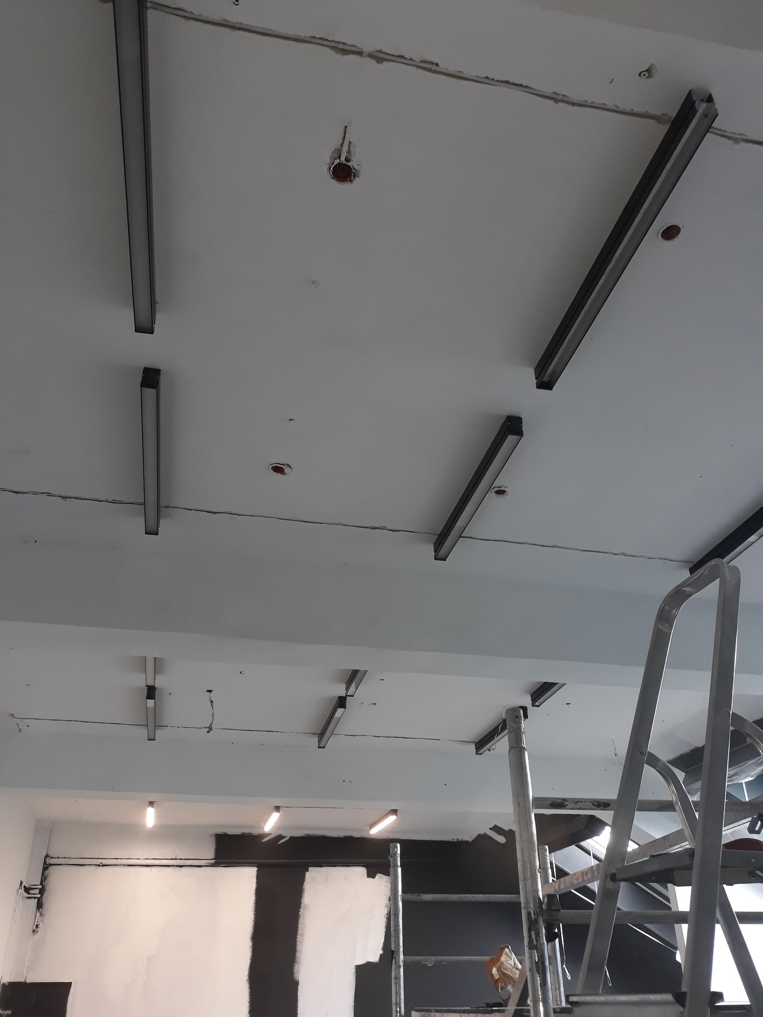 Montaż nowoczesnego oświetlenia liniowego na suficie, widoczne kable i drabina. Prace w toku, sufit w trakcie wykończenia. Ujęcie z dołu, perspektywa z drabiny.