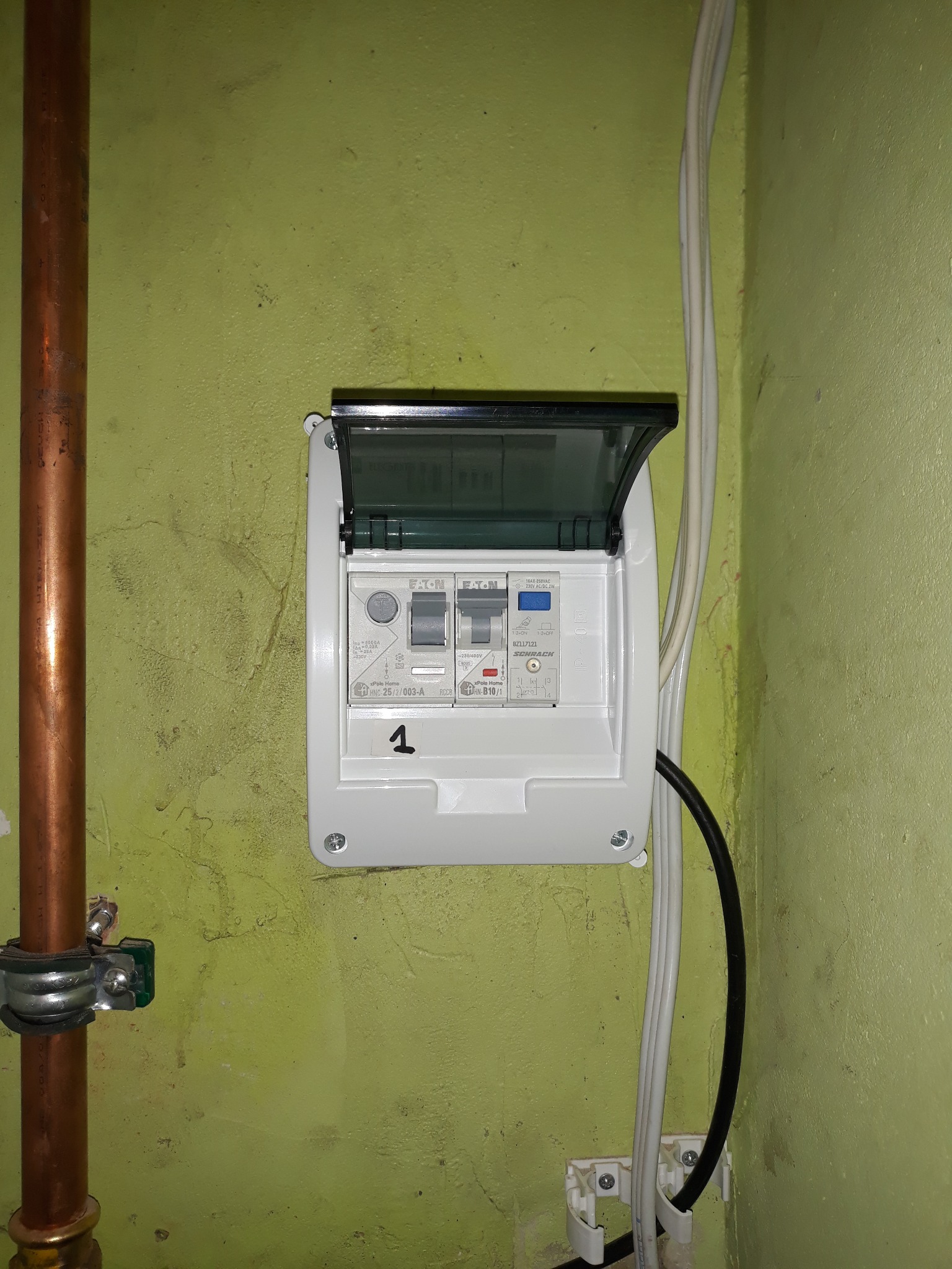 Biała skrzynka elektryczna Eaton z otwartą klapką, zamontowana na zielonej ścianie. Widoczne przewody i rura miedziana. Zasilanie pieca gazowego.