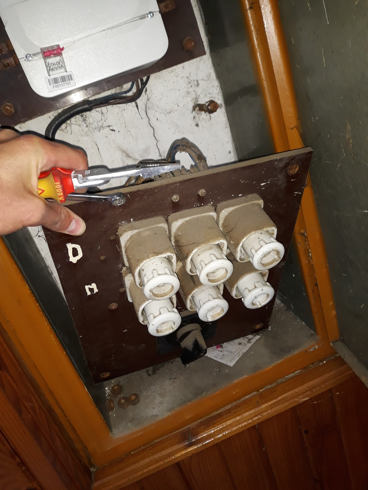Stara tablica bezpiecznikowa typu Dm z ceramicznymi wkładkami topikowymi, częściowo zdemontowana, widoczna ręka elektryka z narzędziem.