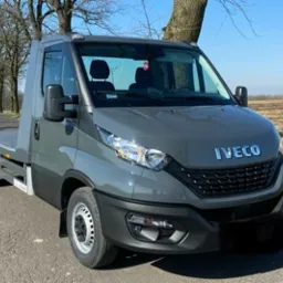 Szara laweta Iveco z platformą załadowczą, zaparkowana na asfaltowej drodze w słoneczny dzień, widok z przodu na tle drzew i pola.