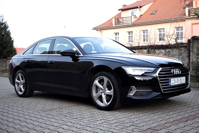 Czarna limuzyna Audi A6 z aluminiowymi felgami, zaparkowana na brukowanym podjeździe przed domem z czerwonym dachem i szarym ogrodzeniem.