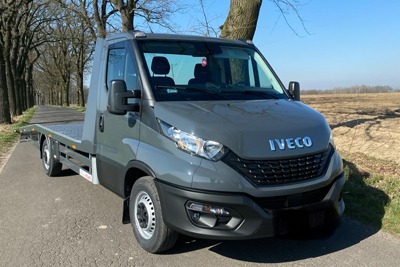 Szara laweta Iveco z platformą załadowczą, zaparkowana na asfaltowej drodze w słoneczny dzień, widok z przodu na tle drzew i pola.