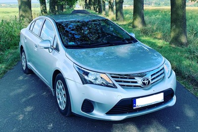 Srebrny samochód osobowy Toyota Avensis na asfaltowej drodze w otoczeniu drzew.