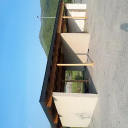 Surowy stan zadaszonej konstrukcji z drewnianą więźbą dachową, jasnymi ścianami i kostką brukową, na tle zielonego wzgórza i błękitnego nieba.