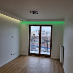 Pusty pokój z drewnianą podłogą, białymi ścianami, grzejnikiem i balkonem z widokiem na miasto, oświetlenie LED w kolorze zielonym nad balkonem i żółtym na ścianie bocznej, nowoczesna lampa sufitowa.