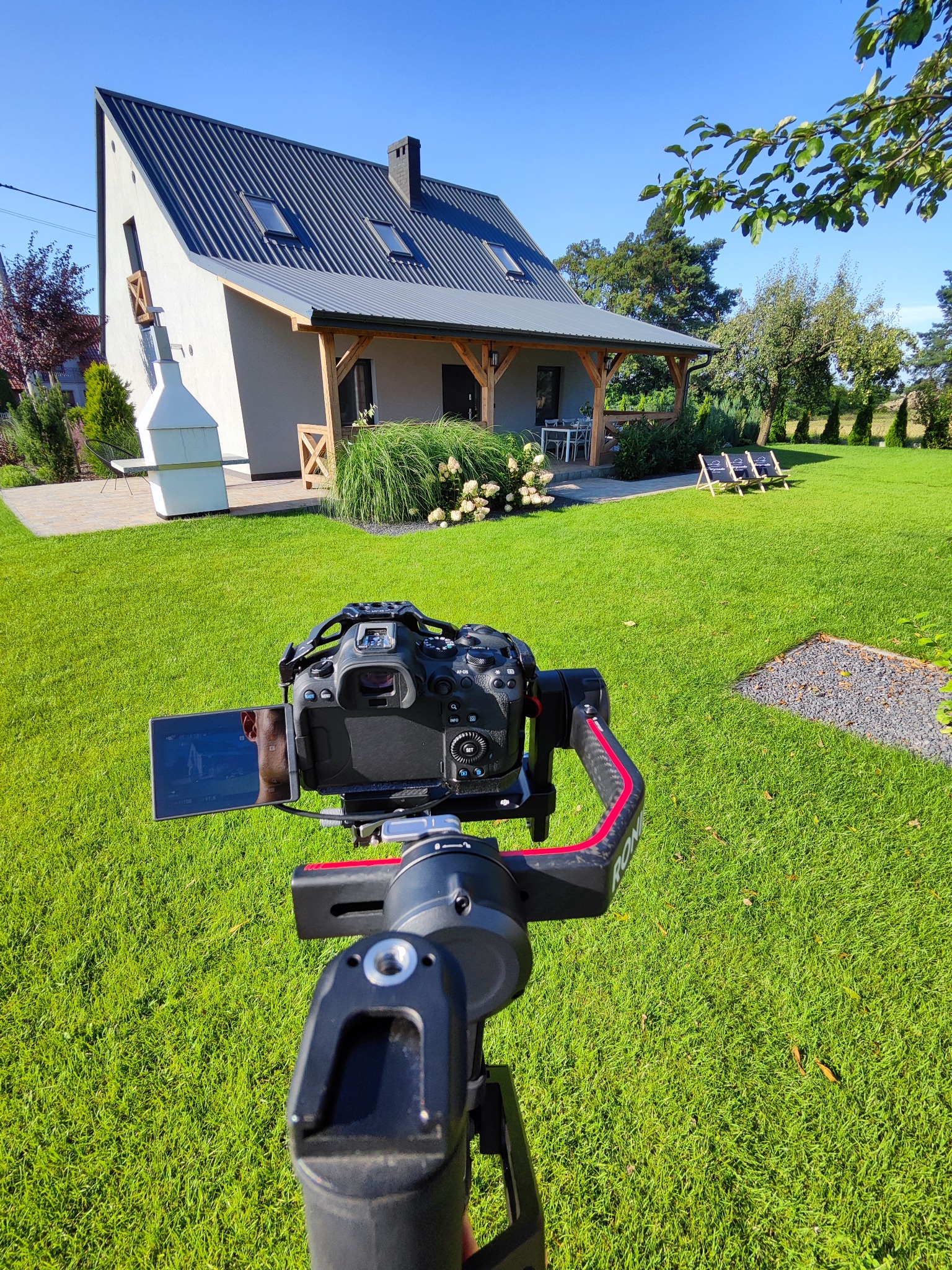 Kamera na gimbalu skierowana na nowoczesny dom z ogrodem w słoneczny dzień, widoczny ekran kamery z odbiciem twarzy.