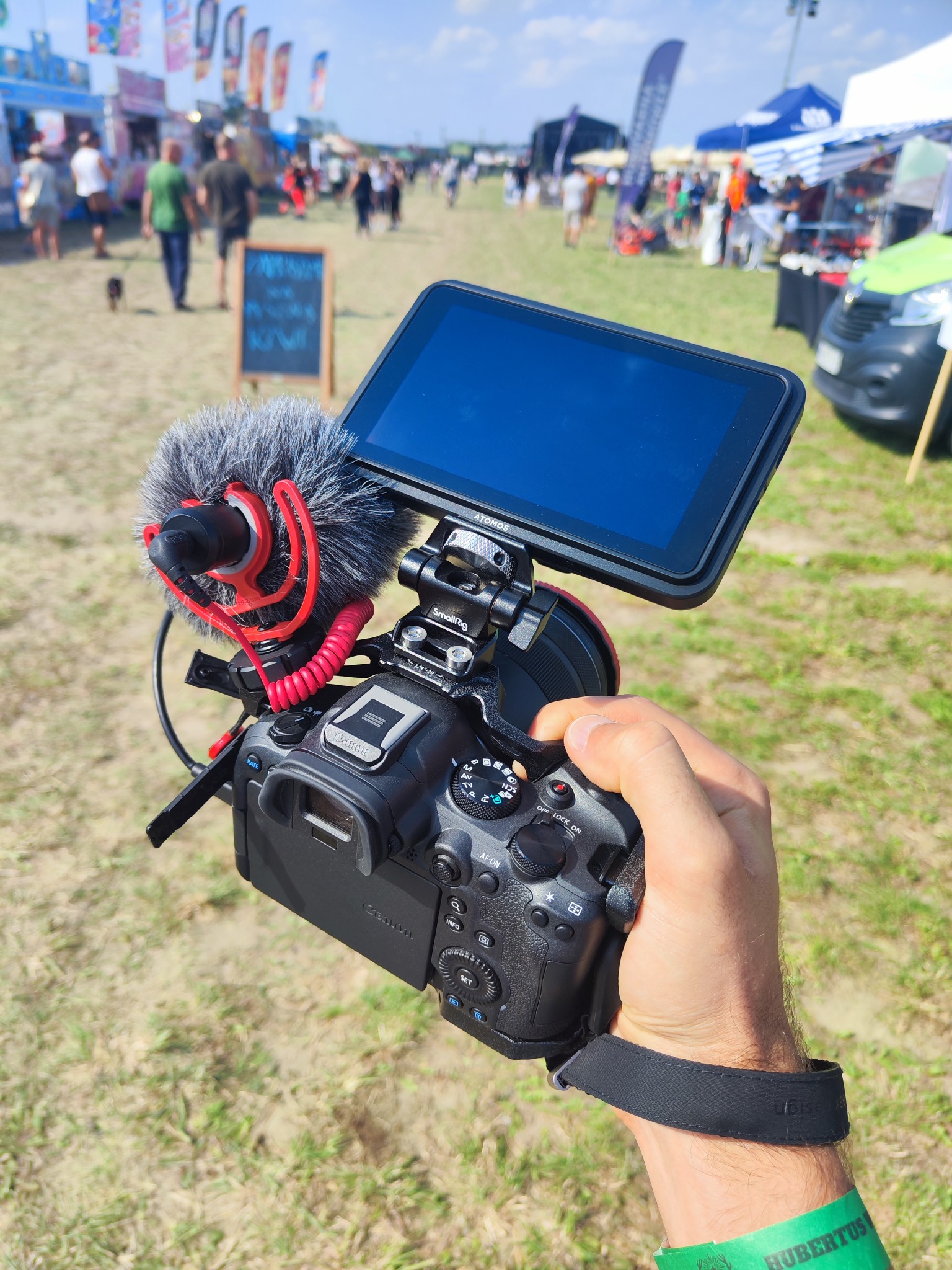 Ręka trzyma aparat Canon z mikrofonem typu shotgun i monitorem podglądowym, w tle plenerowa impreza z straganami i ludźmi.