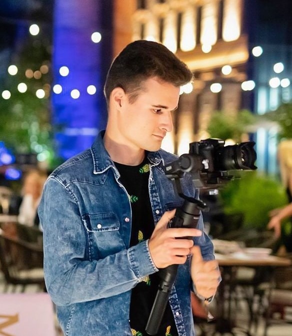 Młody mężczyzna w dżinsowej koszuli trzymający stabilizator z zamontowaną kamerą, ustawiający ostrość w plenerze, w tle oświetlony budynek.