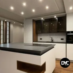 Nowoczesna kuchnia z wyspą i szarym blatem, białe szafki z ciemnymi, przeszklonymi górnymi szafkami, oświetlenie punktowe w suficie, drewniane akcenty i logo firmy w rogu.