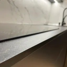 Fragment nowoczesnej kuchni z blatem z ciemnego drewna, płytą indukcyjną i baterią w tle, oświetlenie LED pod szafkami.