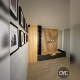 Nowoczesna zabudowa ściany telewizyjnej z drewnianymi lamelami, szafkami w kolorze czarnym i drewnianymi p&oacute;łkami, uzupełniona o czarne ramki ze zdjęciami na ścianie po lewej stronie.