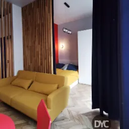 Nowoczesne wnętrze z drewnianą ażurową ścianką działową, żółtą sofą i widokiem na sypialnię z żółtą pościelą, parkiet w jodełkę.