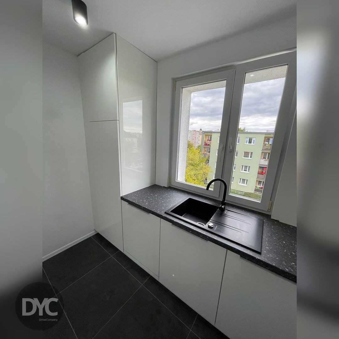 Nowoczesna kuchnia z białymi szafkami i czarnym blatem z widokiem na blok z okna.