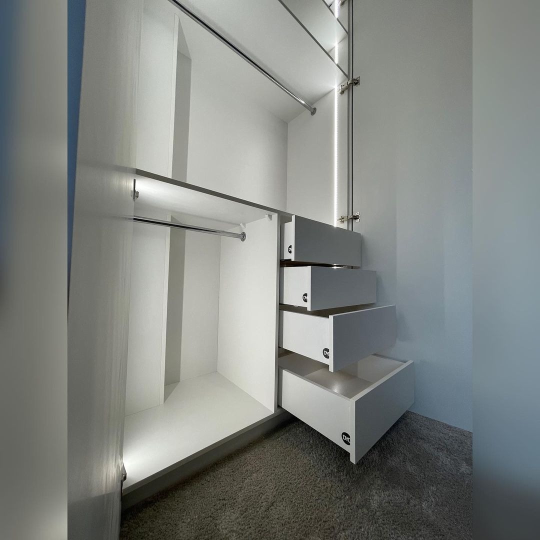 Wnętrze białej szafy z wysuwanymi szufladami, oświetleniem LED i drążkami na ubrania, widoczne logo producenta na frontach szuflad.