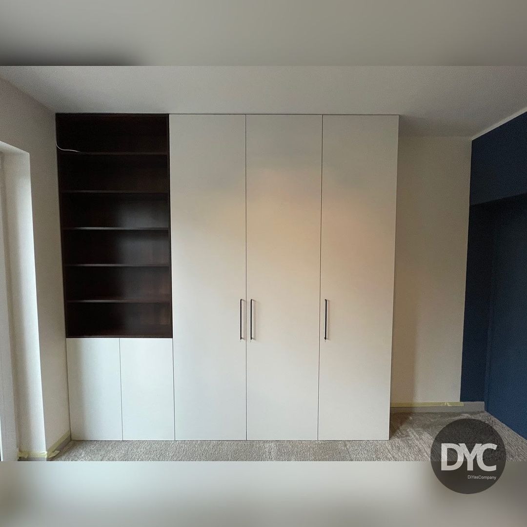 Biała szafa wnękowa z czterema drzwiami i czarnymi, minimalistycznymi uchwytami, przylegająca do otwartej półki z ciemnego drewna, na tle szarej wykładziny.